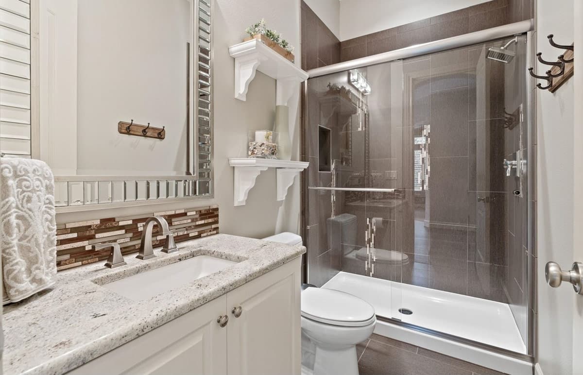 Interior bathroom with vanity, granite countertop, mosaic tile backsplash, toilet, and glass-enclosed tiled shower.