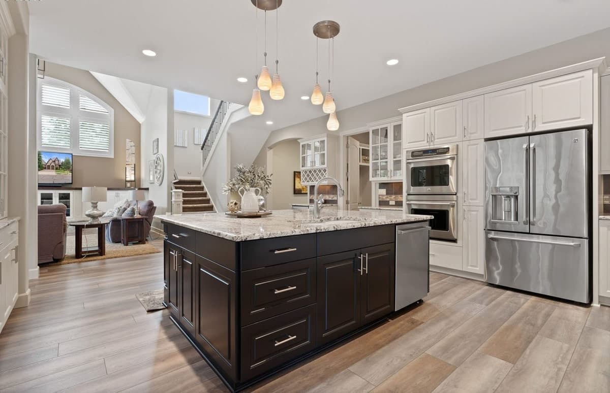 Modern kitchen with dark island, granite counters, stainless steel appliances, and an adjacent open-concept living area with a grand staircase.