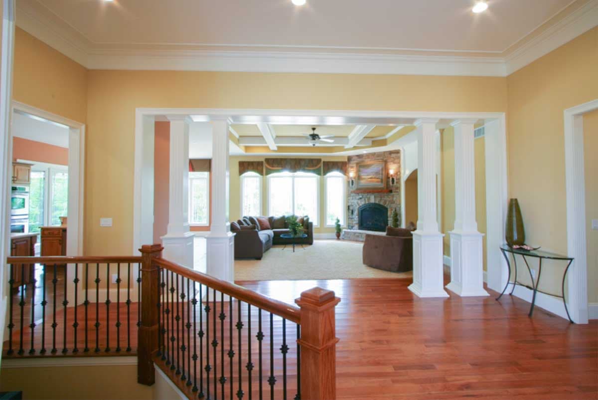 Interior view of foyer with staircase railing and open view to living room with stone fireplace and coffered ceiling.