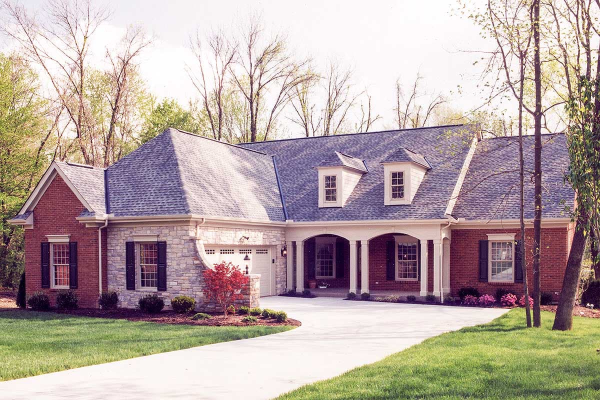 House plan exterior. One-story brick and stone house with dormers, a columned porch, and a side-entry garage.