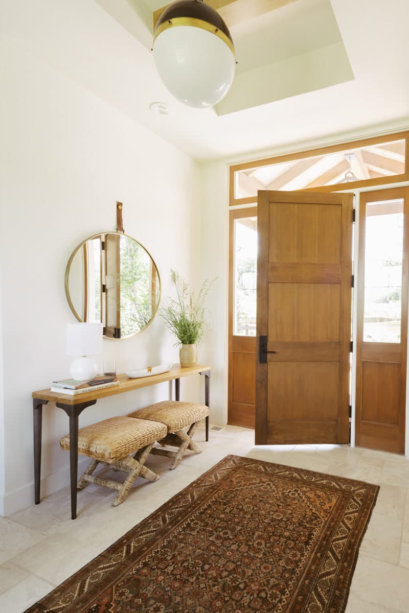 Interior view: Entryway with double wood doors, console table, two woven stools, round mirror, and patterned rug.