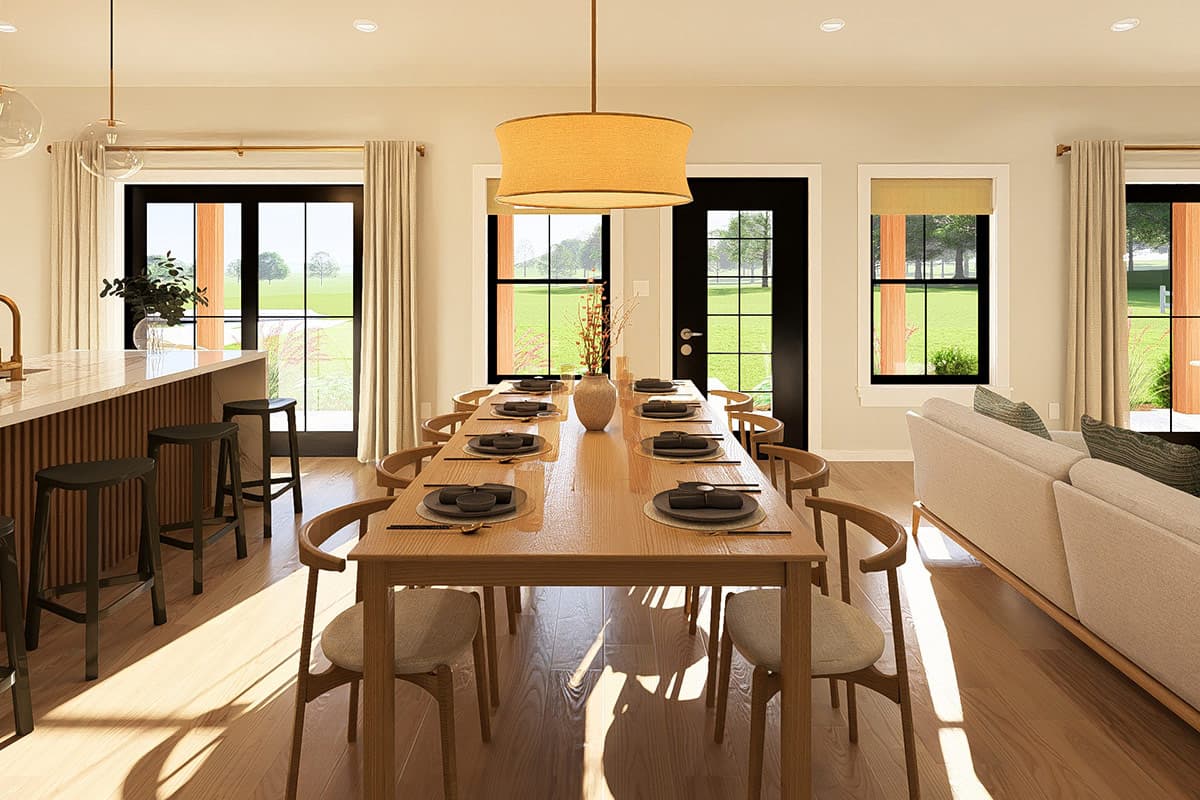 Dining area with a long table set for eight, island with bar stools, and large windows overlooking a lawn.