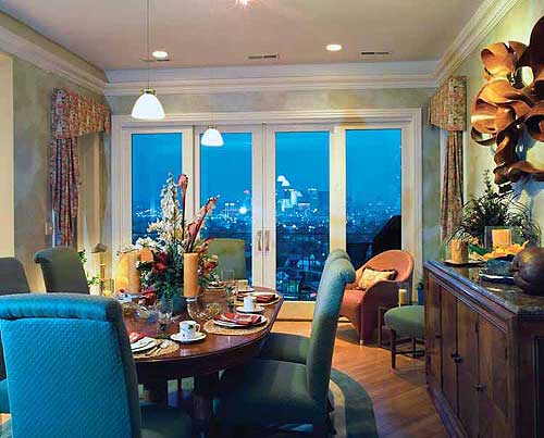 Interior view of a dining room with a large oval table, chairs, and sliding glass doors opening to a city view.