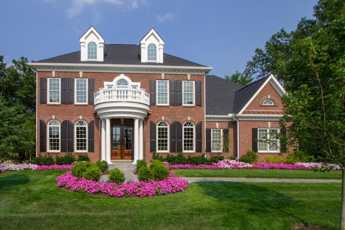 House plan exterior. Two-story brick home with a portico, balcony, and gabled roof. Features include shuttered windows and dormers.