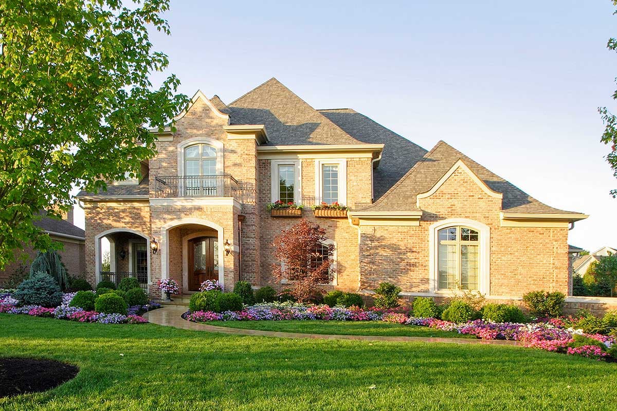 House plan exterior with brick facade, arched entryway, second-story balcony with decorative railing, and multiple gabled rooflines.
