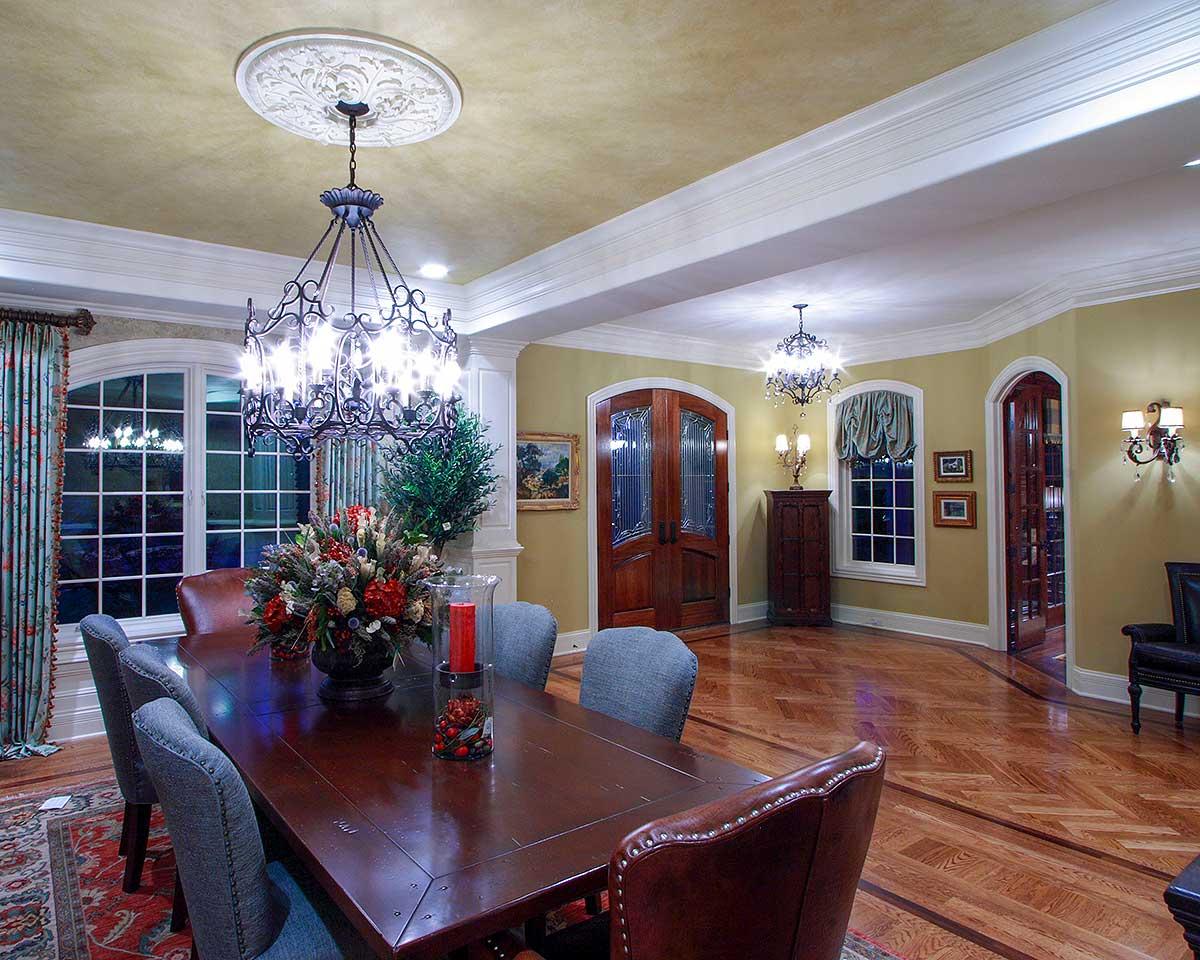 Dining room interior with a large wooden table, upholstered chairs, arched windows, double doors, and ornate chandeliers.