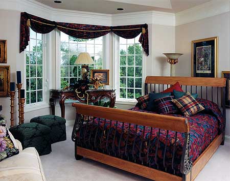 Bedroom with bay windows, wooden sleigh bed with patterned bedding, and ornate table.