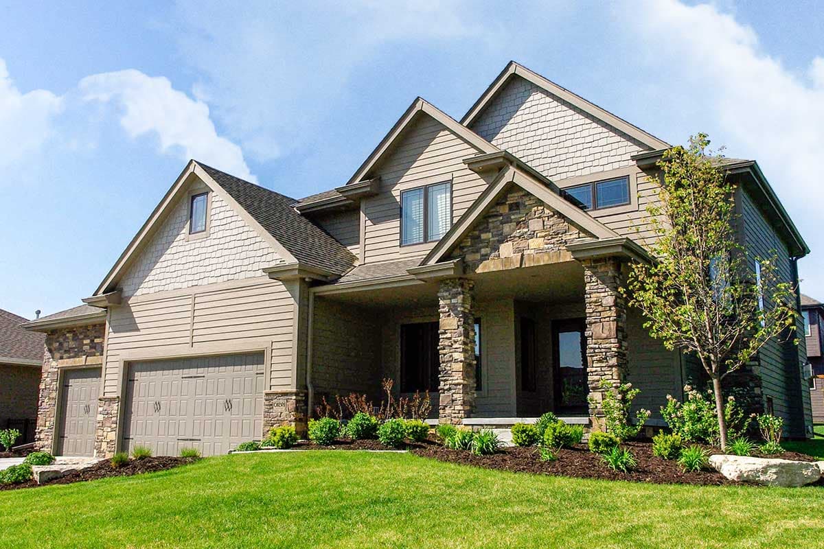 House plan exterior: Two-story home with a covered porch supported by stone pillars, shingle siding, and a triple-car garage.