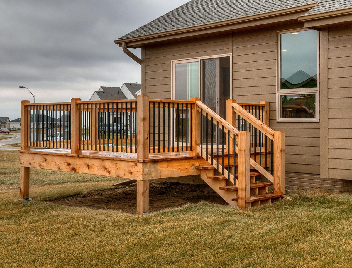 Wooden deck with black metal balusters and stairs attached to a house with horizontal siding and large windows.