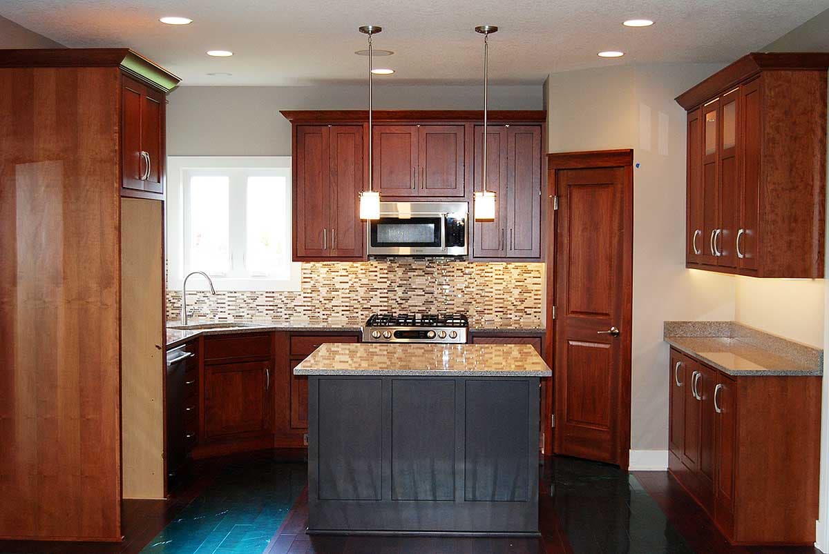 Kitchen interior with cherry wood cabinets, granite countertops, mosaic tile backsplash, gas range, and island.