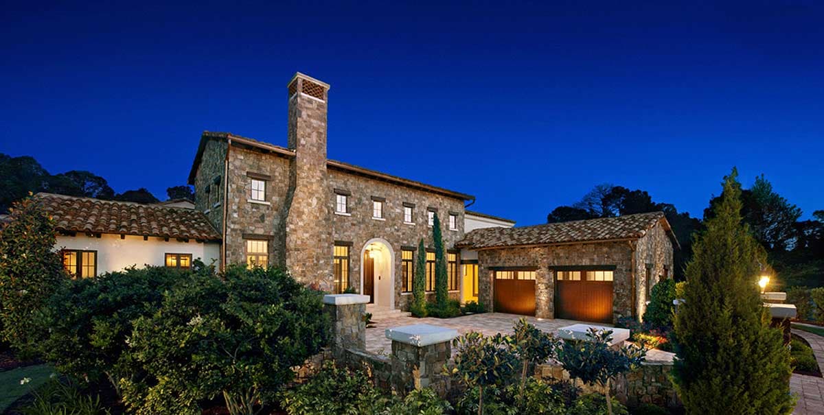 House plan exterior featuring stone facade, multiple gables, arched entryway, and attached two-car garage with wood doors.
