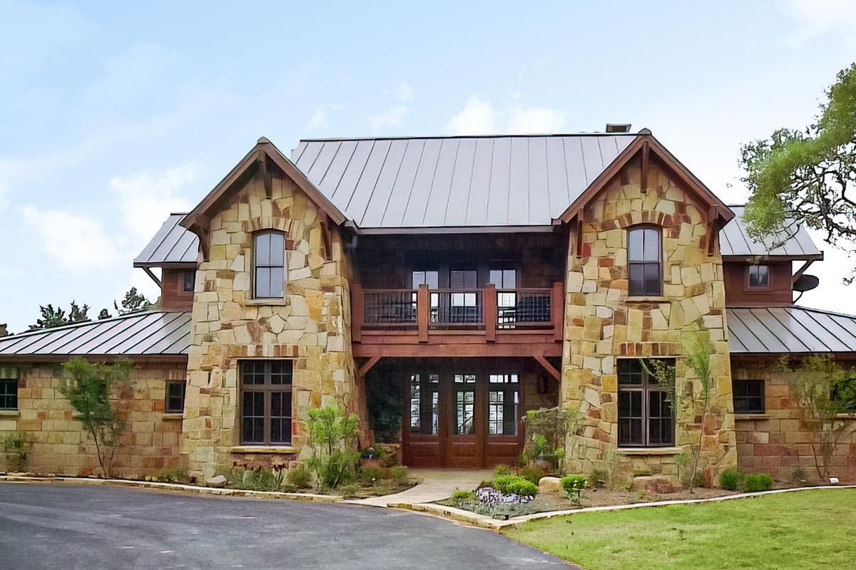 House plan exterior: Two-story stone home with metal roof, prominent gables, second-story balcony, and double entry doors.