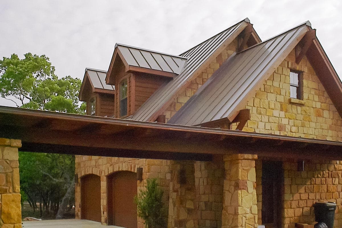 House plan exterior featuring stone walls, metal roofs, gables, dormer windows, and a porte-cochère with arched garage bays.