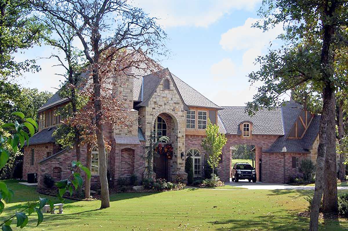 House plan exterior: Two-story brick home with stone accents, arched entryway, multiple gables, and porte-cochère with a vehicle.