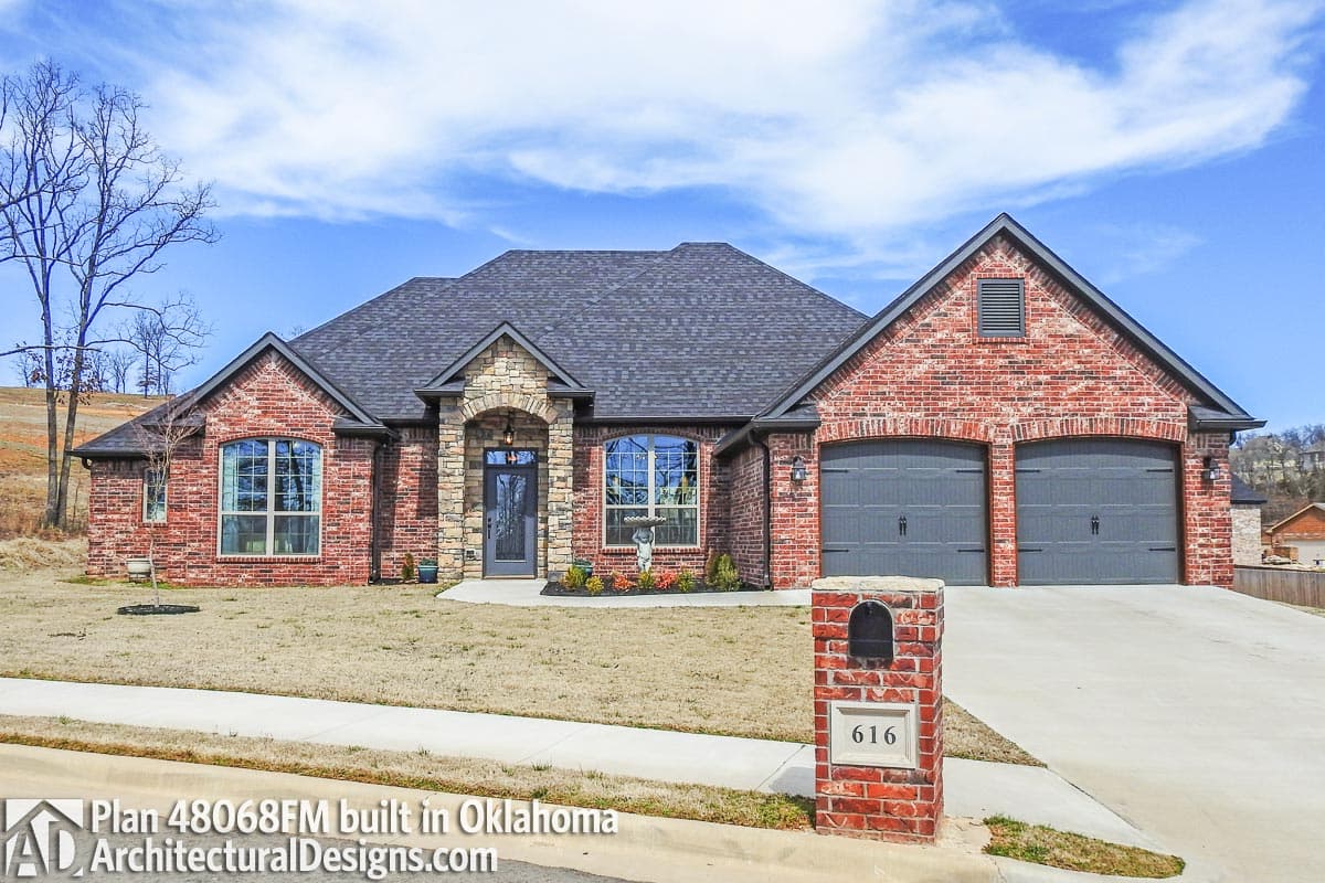 House plan exterior. One-story brick home with gabled roof, stone archway, and attached two-car garage. Features include dormers and arched window.