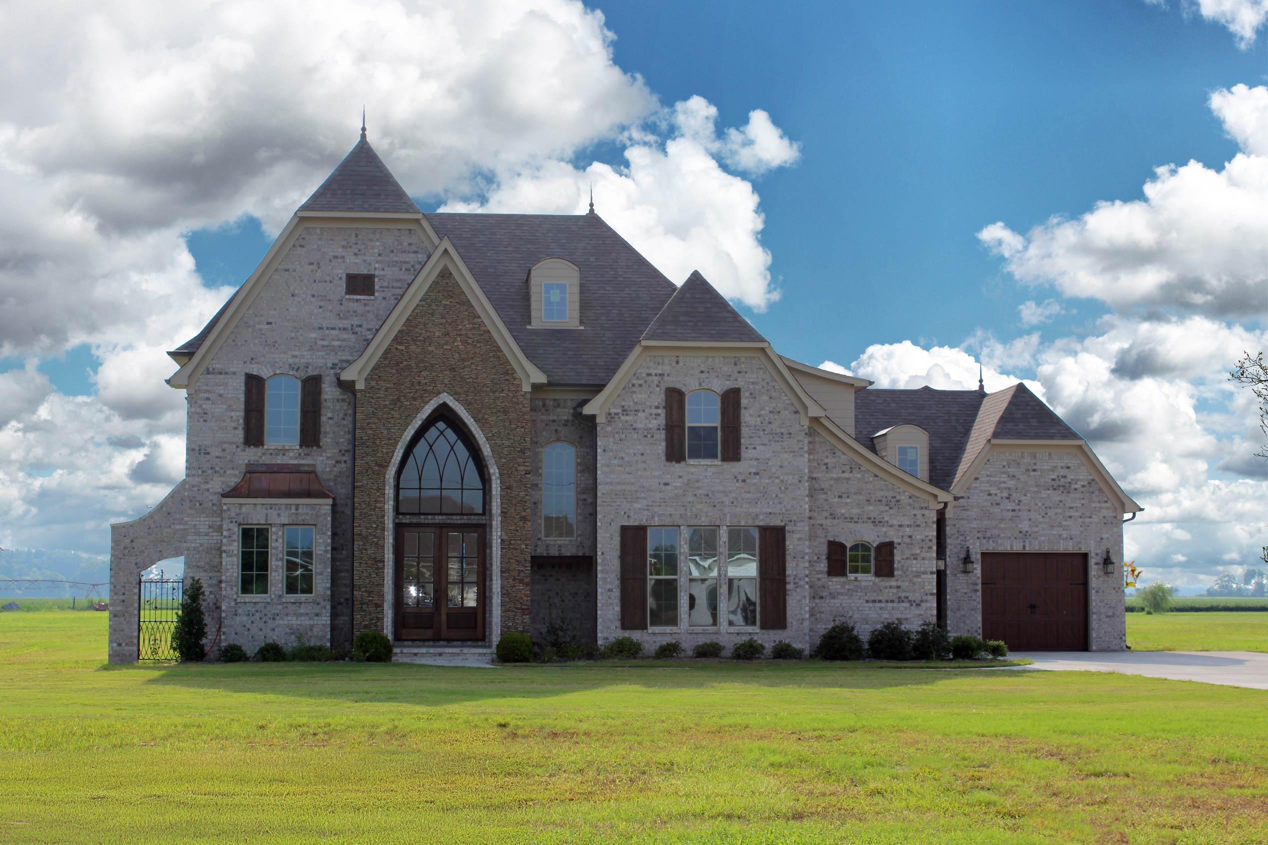 House plan exterior: Two-story brick home with stone accents, arched entryway, multiple gables, dormer windows, and a side-entry garage.