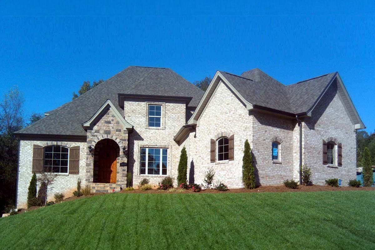 House plan exterior: Two-story home with arched stone entry, light brick facade, multiple gables, and varied window styles.