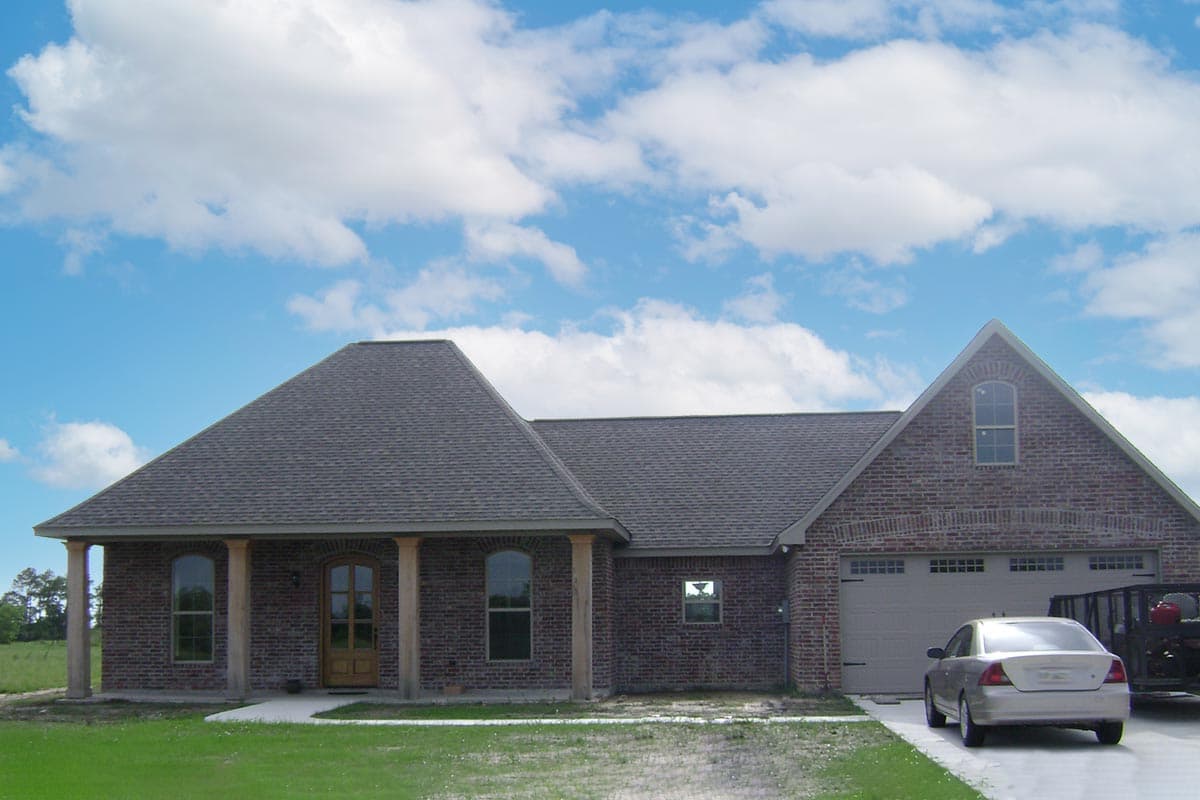 House plan exterior: one-story brick home with hip roof, covered front porch supported by columns, and a side-facing garage.