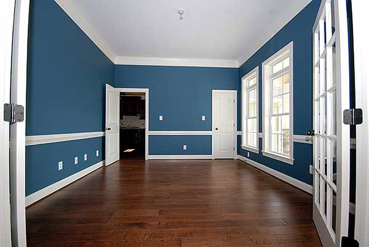 Interior view of an empty room with blue walls, wainscoting, three tall windows, and two doors opening to a kitchen and another room.