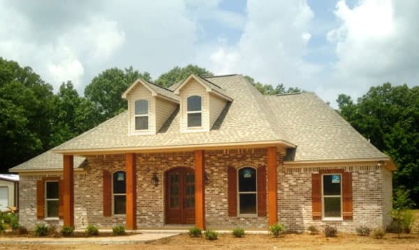 House plan exterior: one-story brick home with a covered front porch supported by wood columns, dormer windows, and a gable roof.