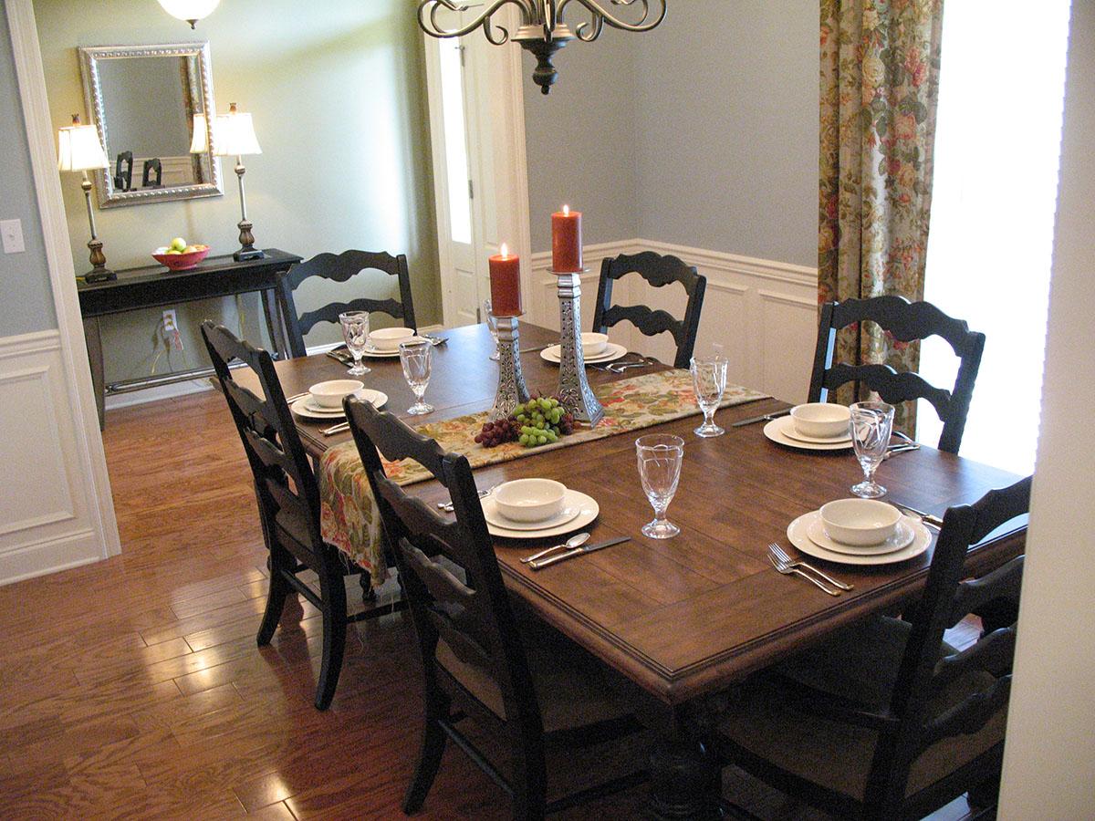 Dining room with wooden table set for eight, featuring a floral runner, two candlesticks, and dark wood chairs.