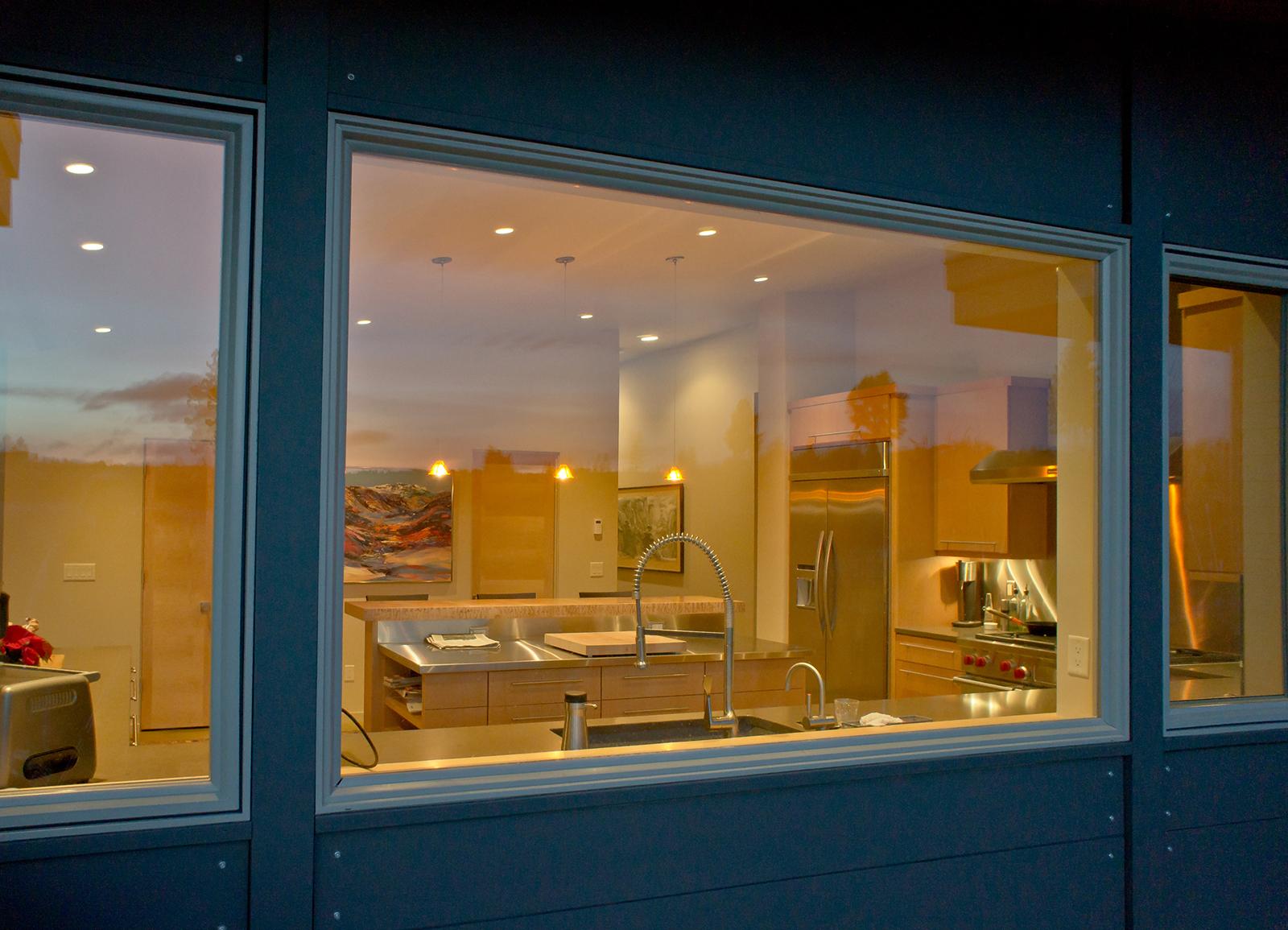 Modern kitchen with stainless steel island, sink, faucet, range, and refrigerator visible through a large window.