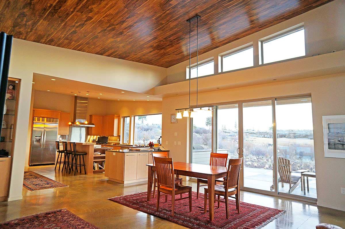 Modern open-plan living area with vaulted wood ceiling, kitchen island, dining table, and large windows.