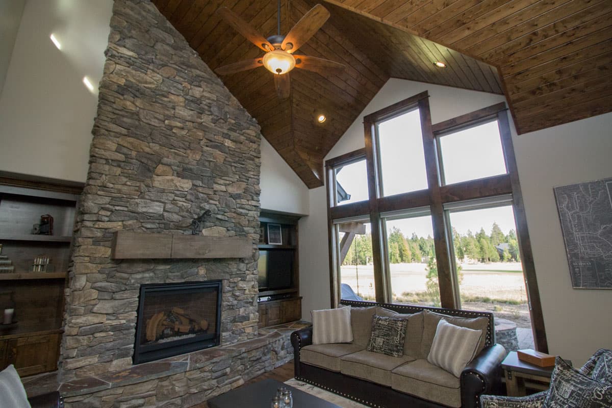 Interior view of a living room with a floor-to-ceiling stone fireplace, vaulted wood ceiling, and large windows.