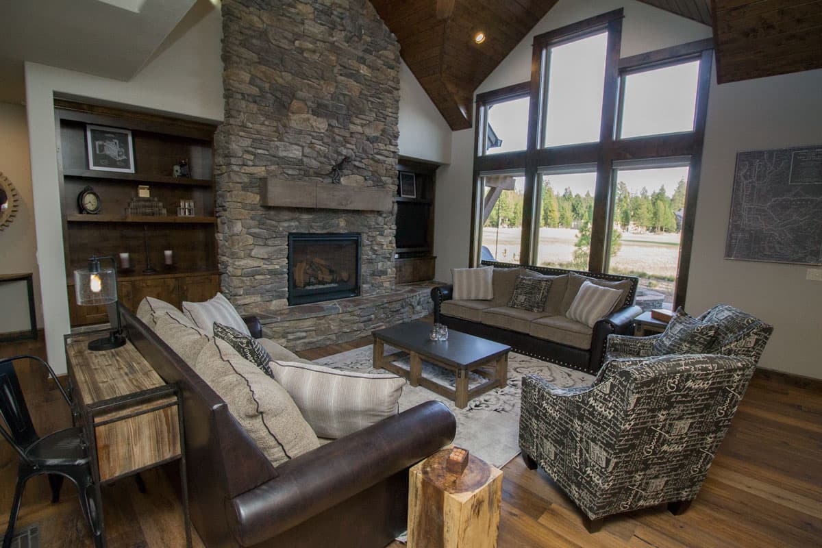Living room with stone fireplace, vaulted wood ceiling, built-in shelving, and large windows overlooking a wooded landscape.