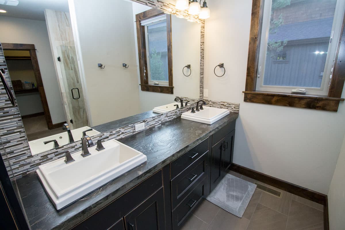 Bathroom with double vanity, dark wood cabinetry, tiled backsplash, and walk-in shower visible in mirror.