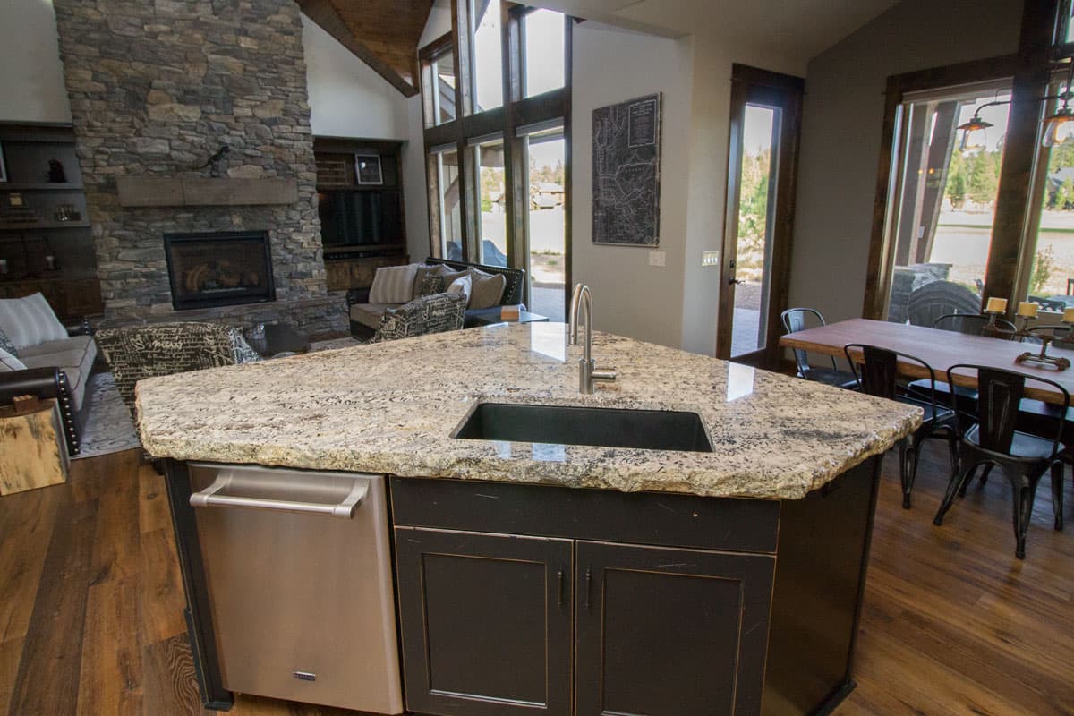 Kitchen island with sink and dishwasher, view towards great room with stone fireplace and dining area.