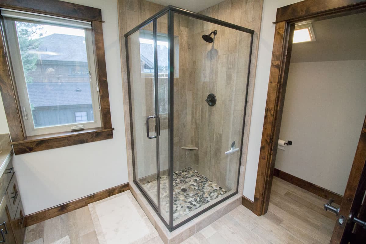 Interior view of bathroom with glass-enclosed shower, tiled walls, pebble floor, and window.