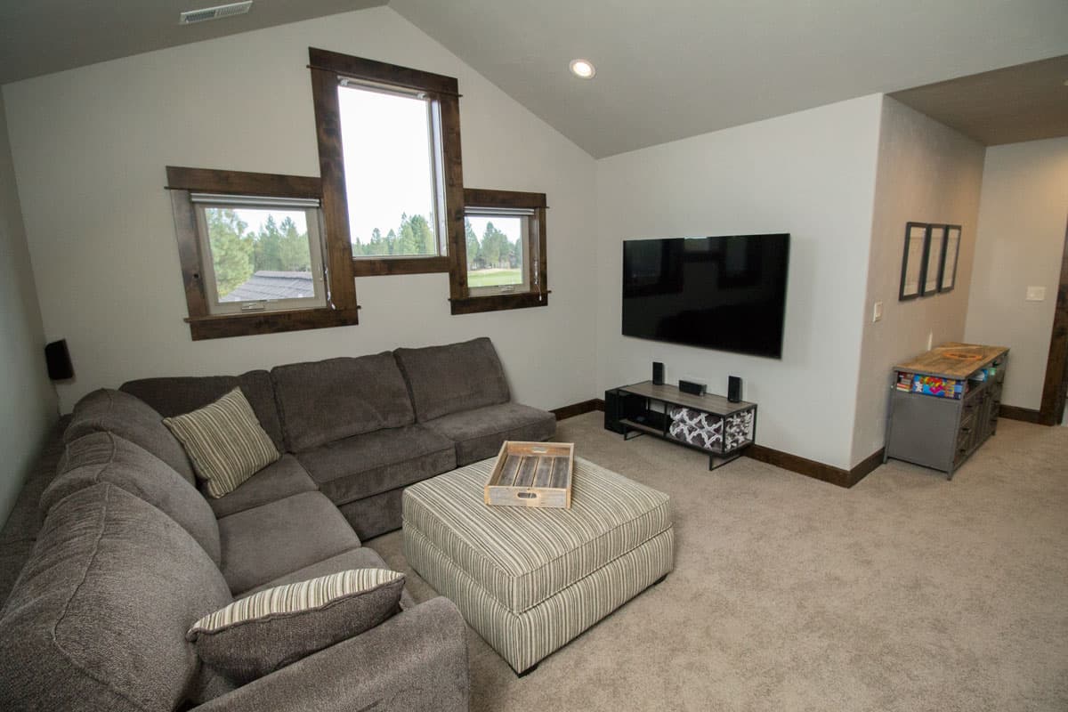 Living room with sectional sofa, ottoman, and mounted television. Sloped ceiling with unique wood-trimmed windows.