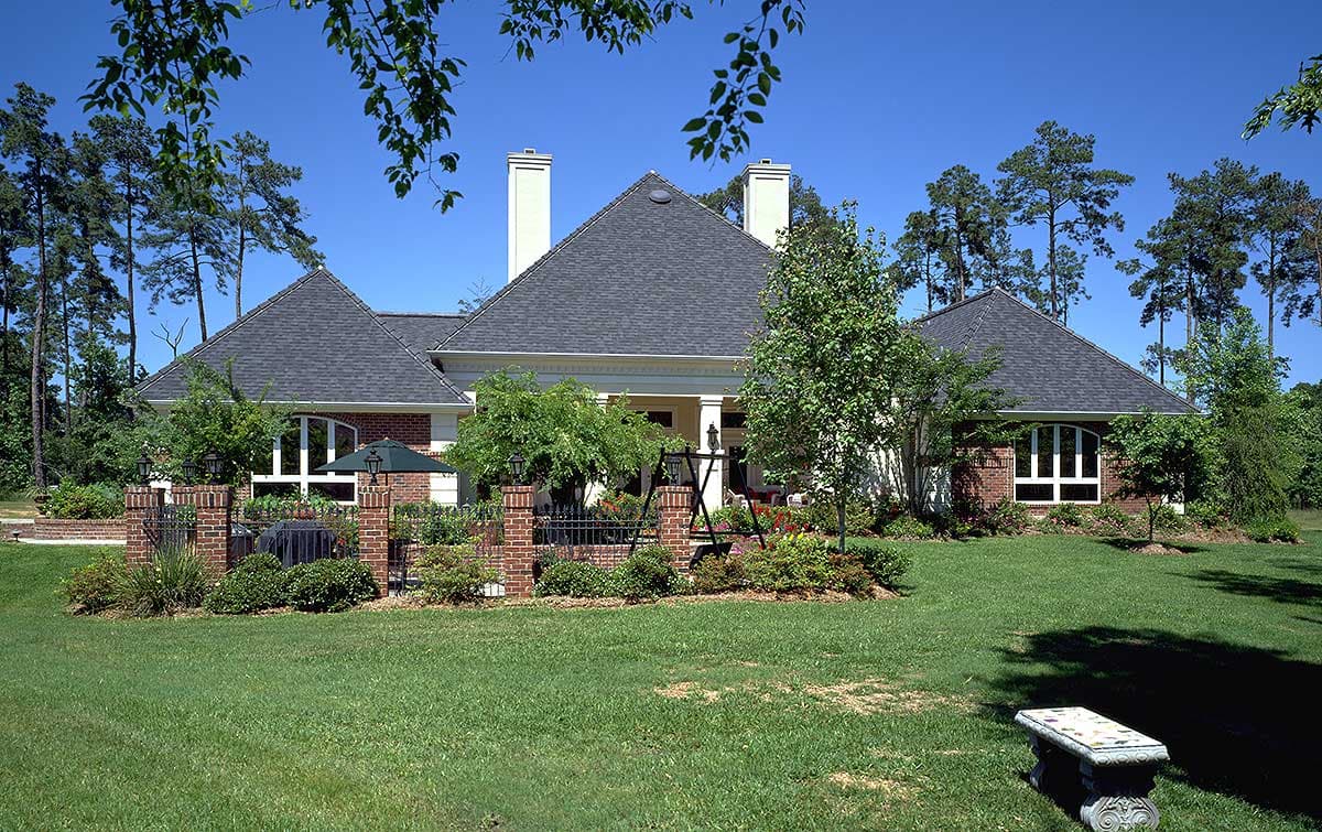 House plan exterior with a dark shingle roof, two chimneys, arched windows, and a brick fence surrounding a patio area.