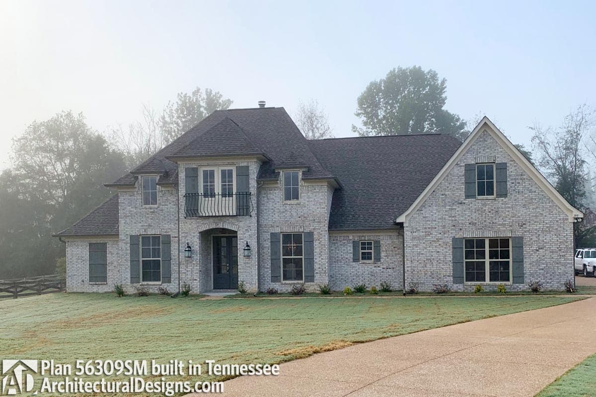 Two-story house plan exterior with white brick, dark shutters, arched entryway, balcony, and multiple gables.