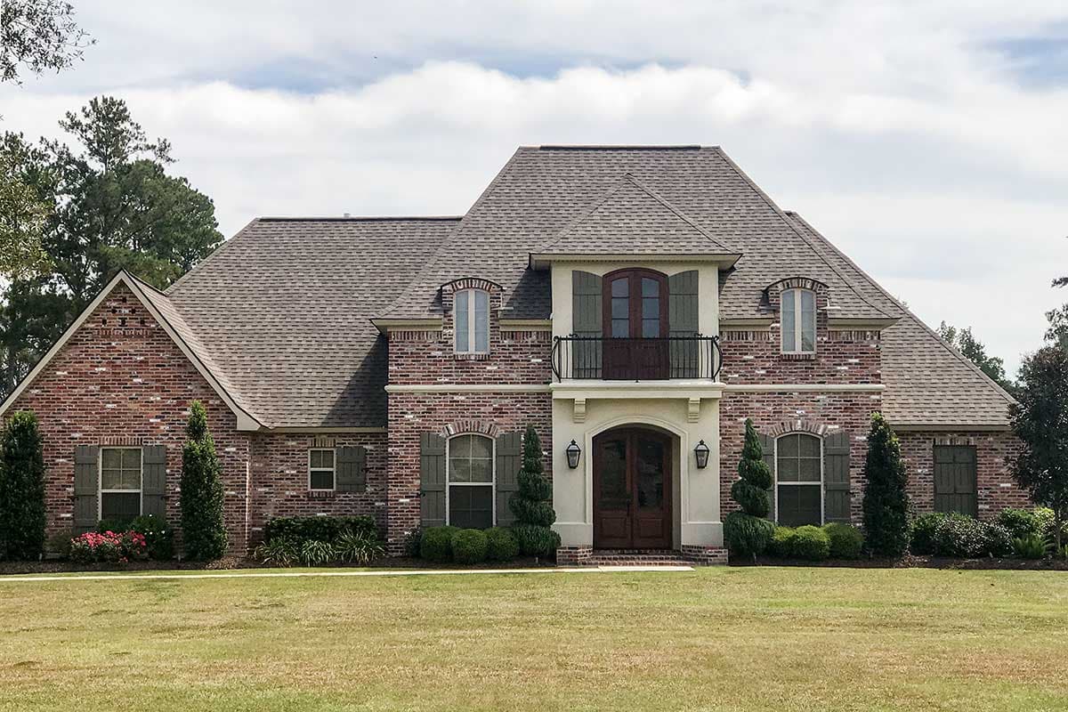 House plan exterior. Two-story brick home with a gable roof, arched windows, shutters, and a balcony over the front entry.