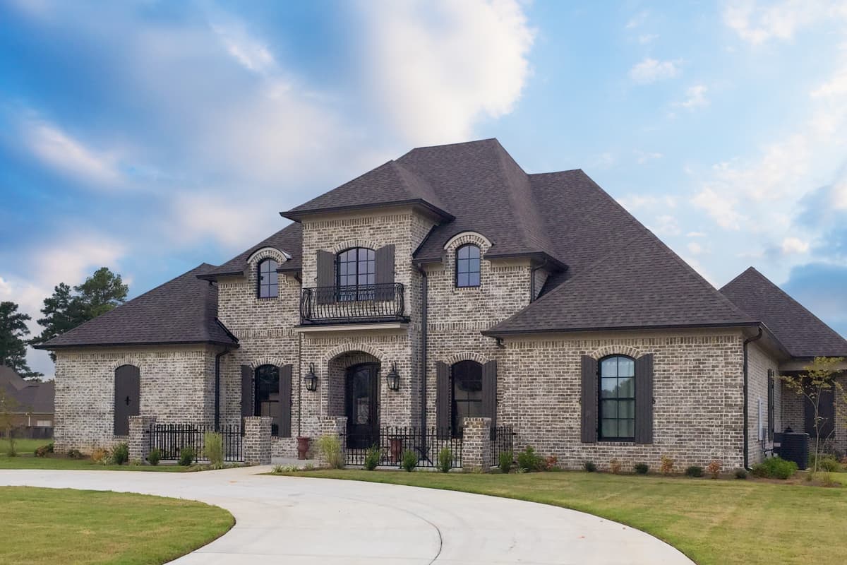 House plan exterior. Two-story brick home with gabled roof, dormers, and arched windows. Includes a wrought-iron balcony and shutters.