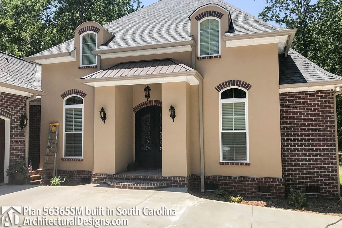 House plan exterior with stucco and brick, arched dormers, metal roof porch, and arched entry.