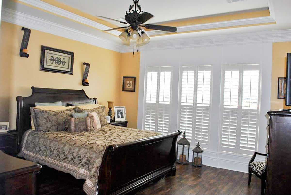 Primary bedroom with sleigh bed, tray ceiling, ceiling fan, and three plantation shutters on windows.