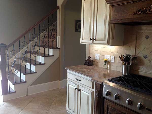 Kitchen with cream cabinets, granite countertop, tiled backsplash, and a staircase with wrought iron railing.