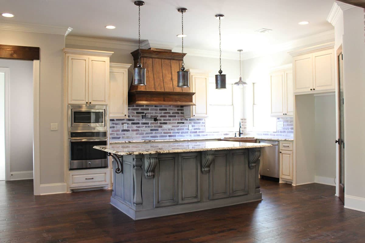 Kitchen with gray island, white cabinets, wood range hood, brick backsplash, and granite countertops.