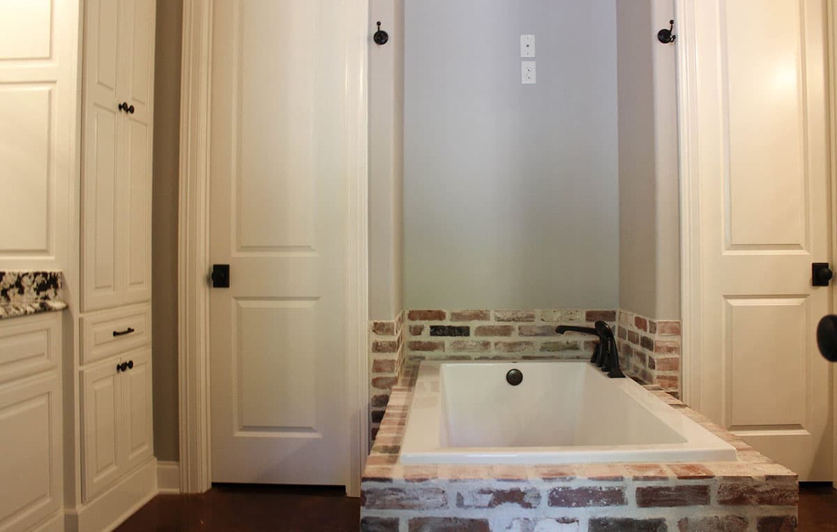 Bathroom interior with a soaking tub surrounded by brick and flanking doors, and tall white cabinetry.