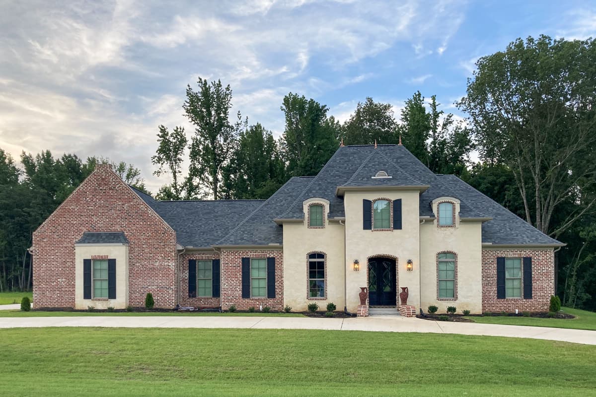 House plan exterior. Two-story, featuring a brick facade, multiple gables, arched entry, and dormers. Black shutters, and dark roof.