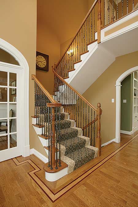 Interior view of a curved staircase with wooden banister, ornate metal balusters, patterned carpet runner, and arched doorway.