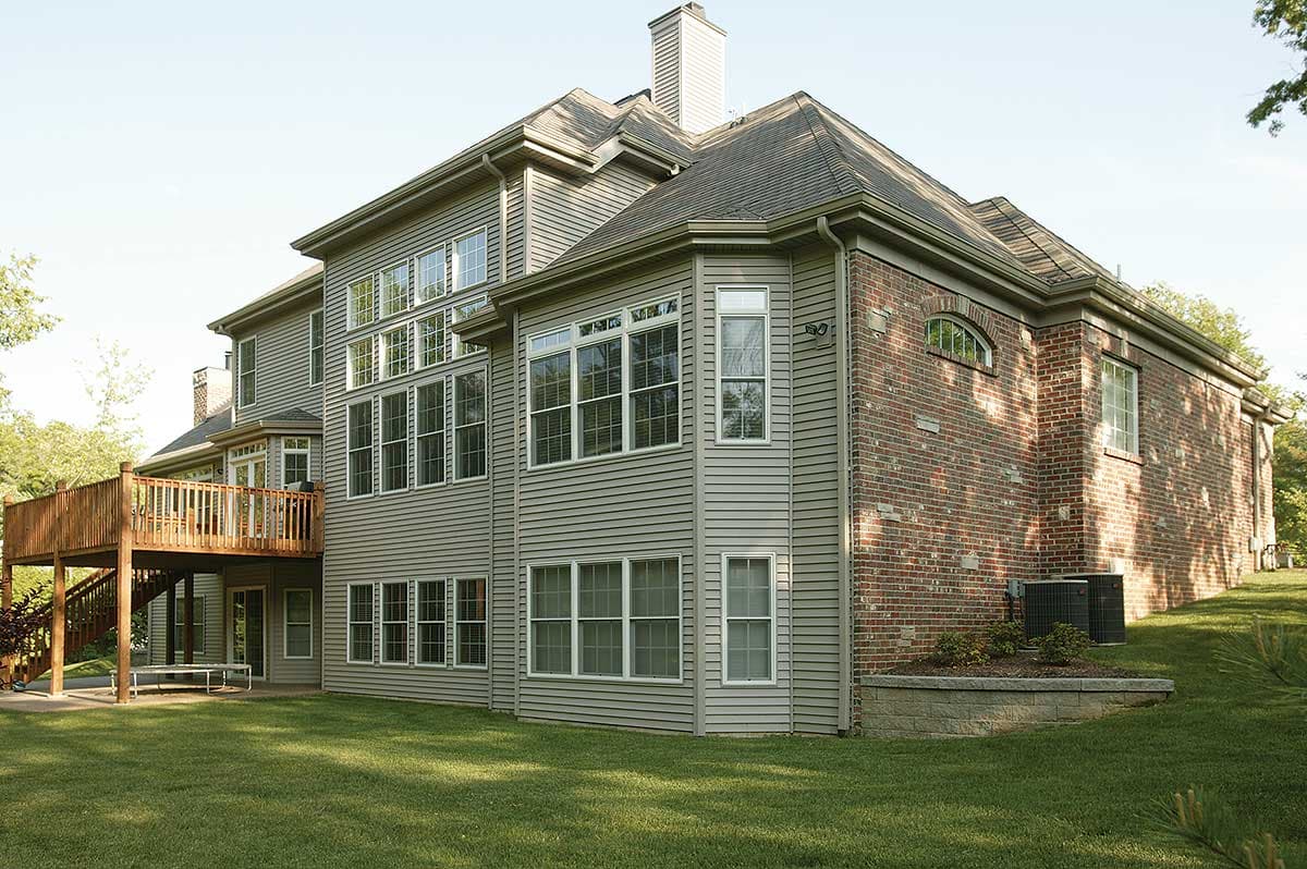 House plan exterior: Two-story home with vinyl siding, brick accents, large multi-pane windows, and a wooden deck.