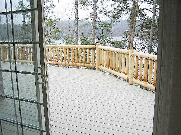 Deck with log railings overlooks a wooded landscape and water. Sliding glass door in foreground.