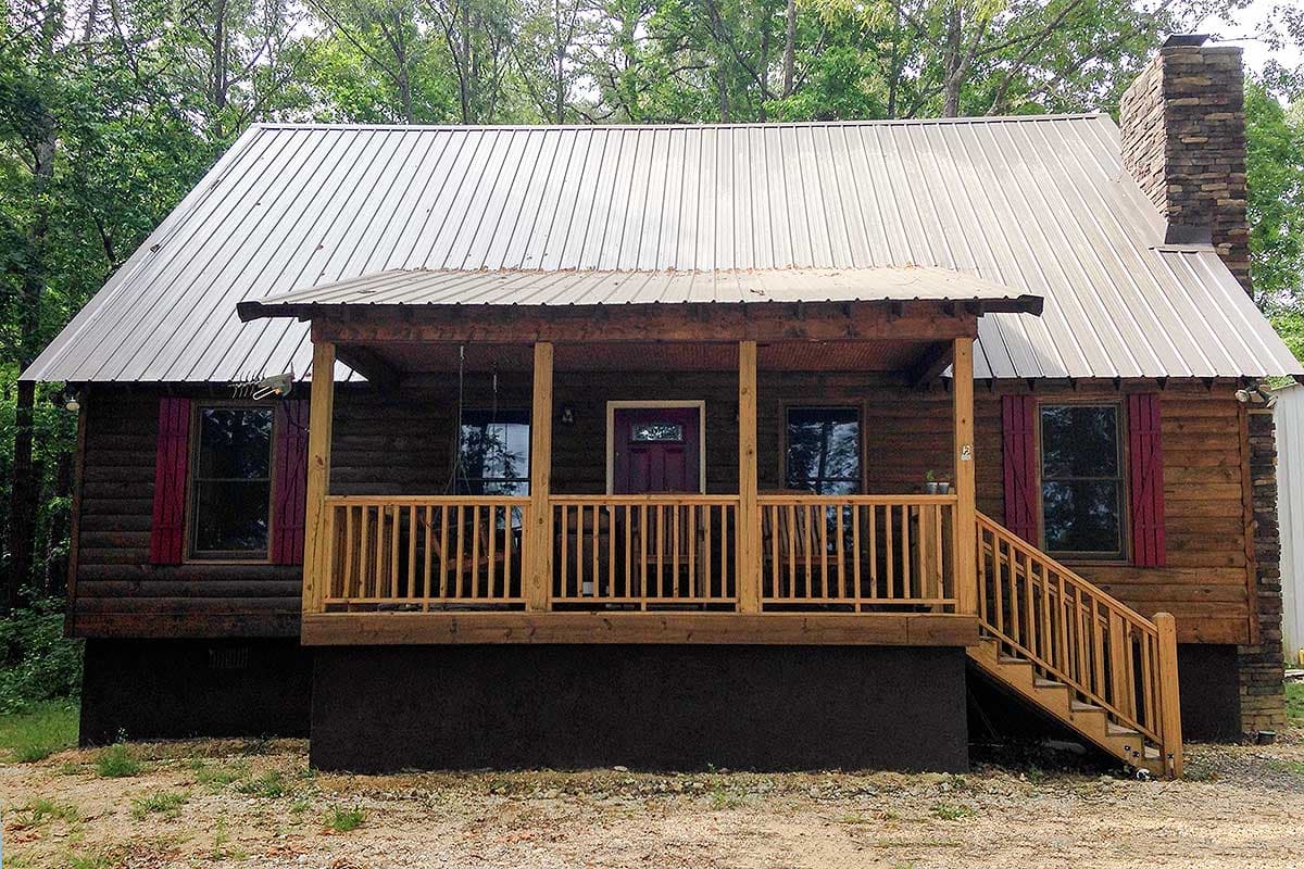 Log cabin exterior with covered porch, gabled roof, stone chimney, and wood shutters. House plan.