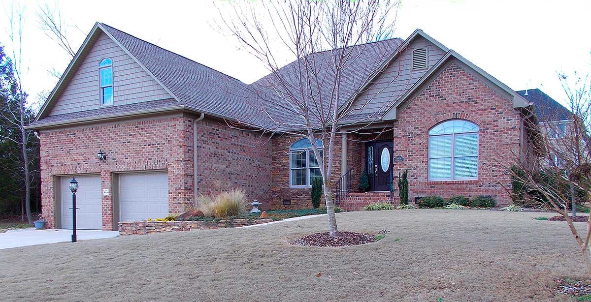 House plan exterior. Two-story brick home with gabled roof, arched windows, and two-car garage. Dormers visible above garage and entry.