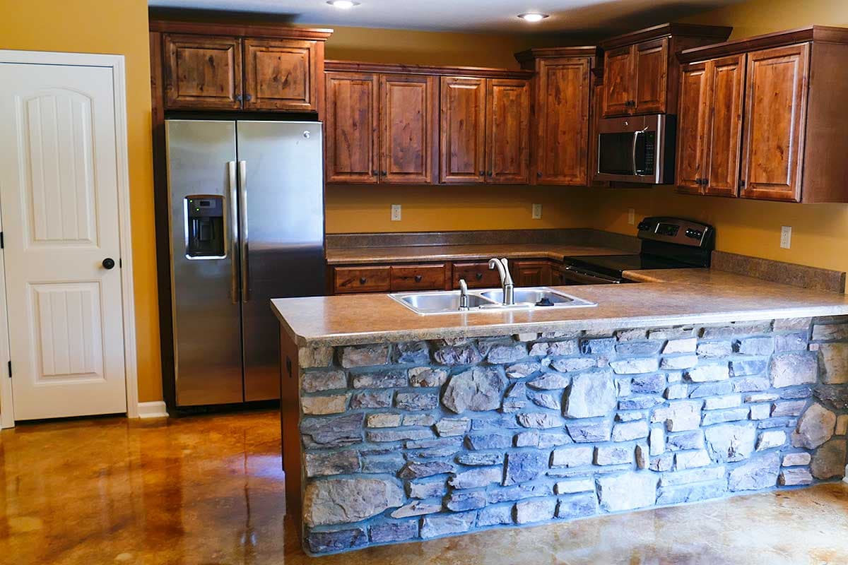 Kitchen interior with stainless steel refrigerator, wooden cabinets, granite countertops, and stone-clad island with sink.