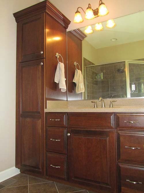 Bathroom vanity with dark wood cabinetry, drawers, and a tall storage cabinet. Mirror reflects tiled shower.